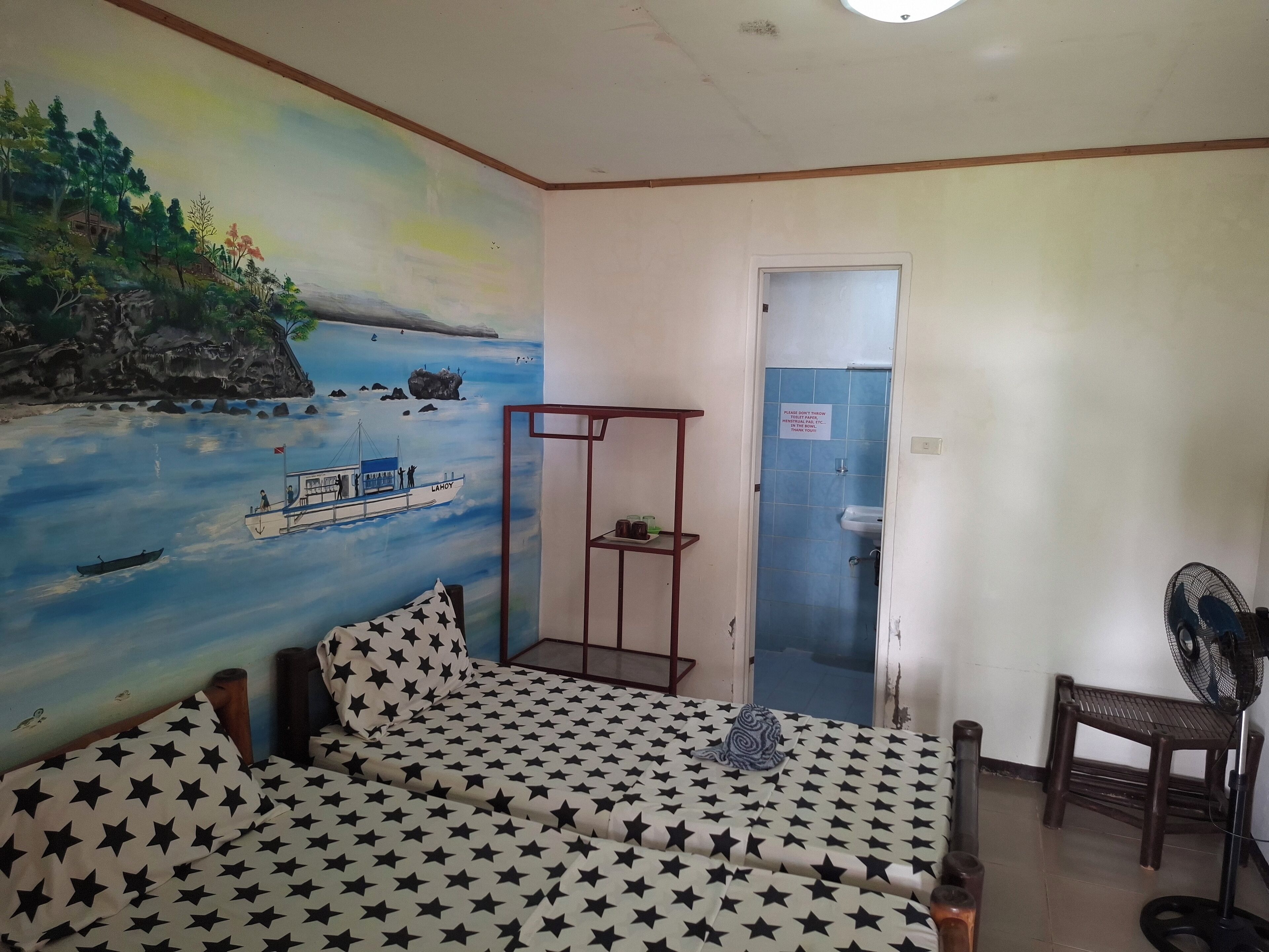 A room with a mural of a coastal scene, a bed with star-patterned bedding, a small shelf, and a fan.