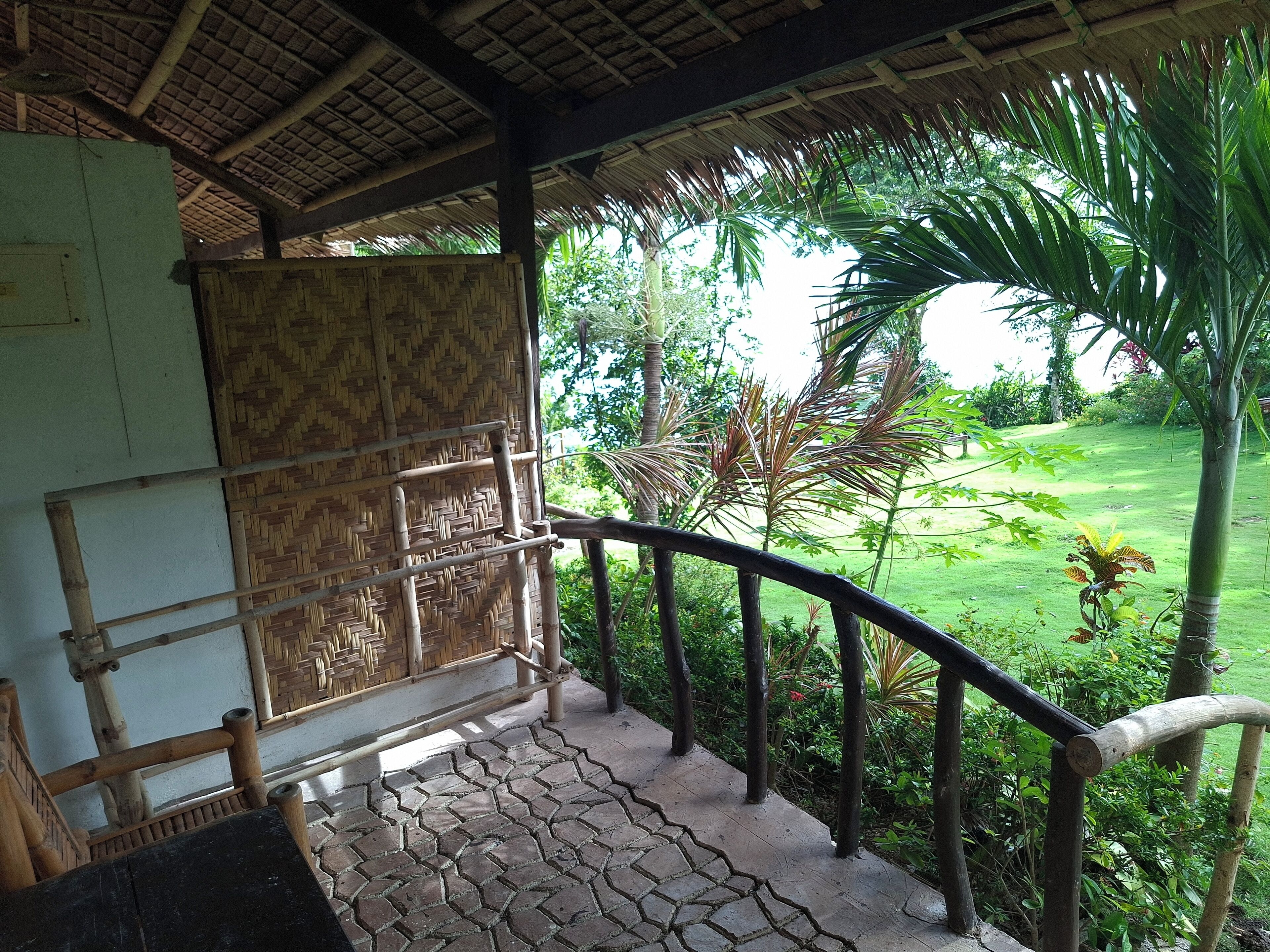 A covered outdoor area with a bamboo railing, a woven wall, and a view of a lush garden.