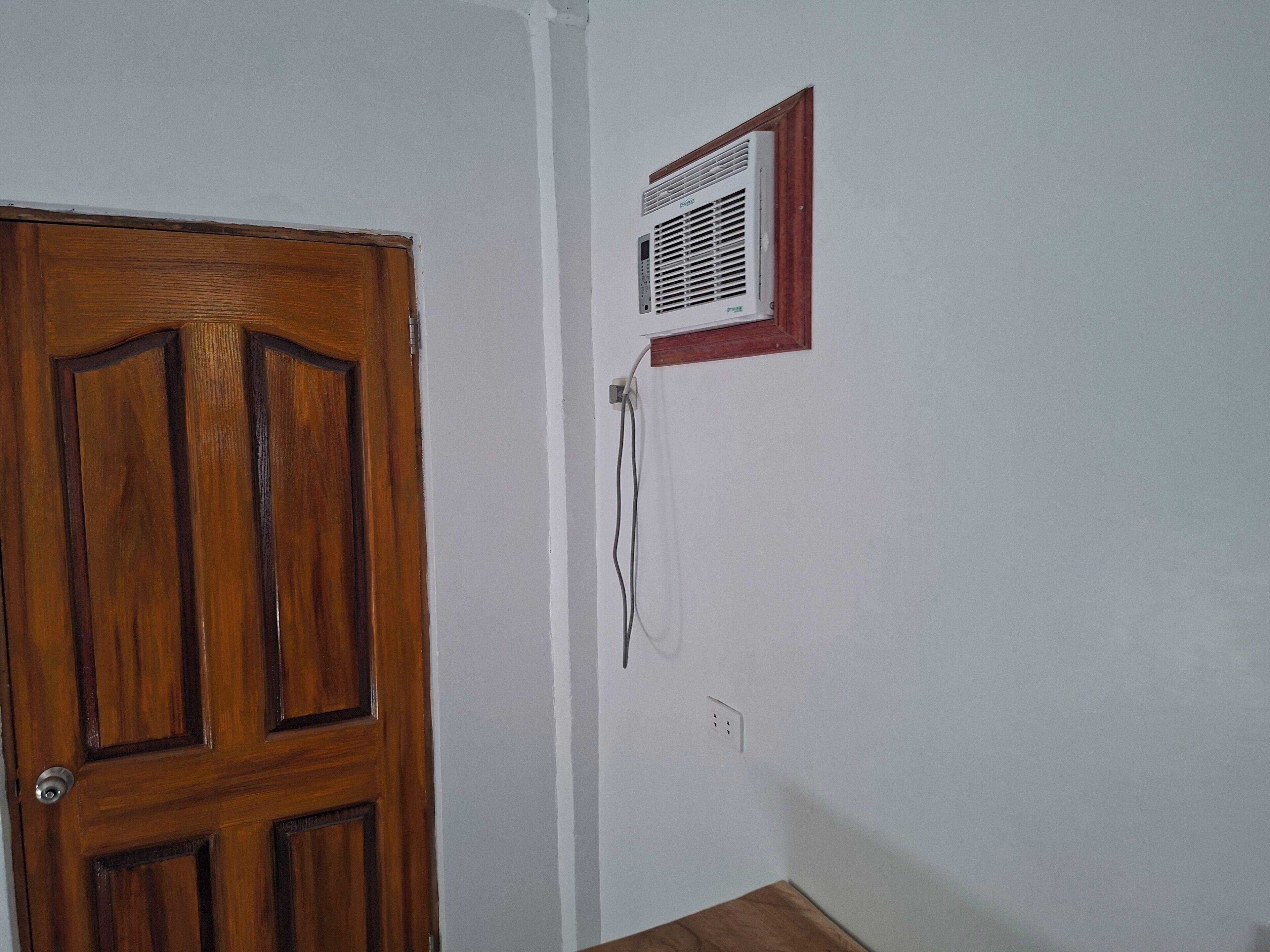 A room with a wooden door, a white wall, and a white air conditioning unit mounted on the wall.