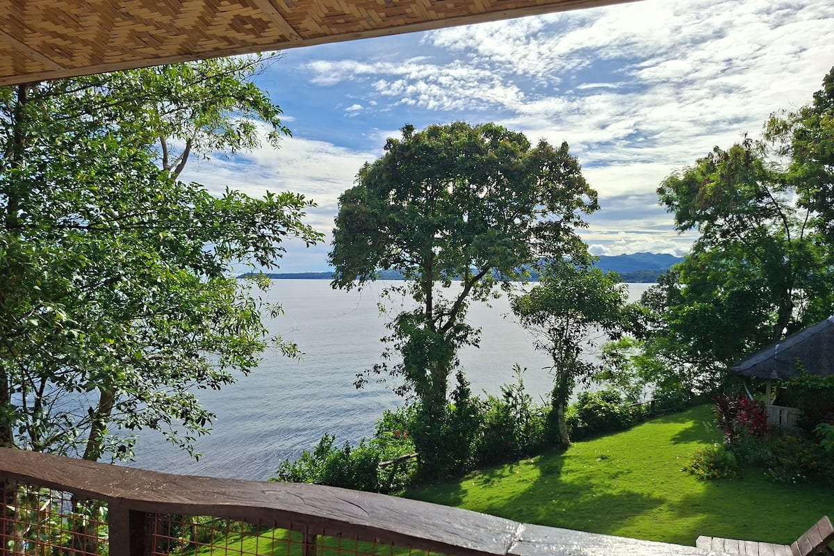 Une vue depuis un espace extérieur abrité, donnant sur un lac bordé d’arbres et d’une zone herbeuse.