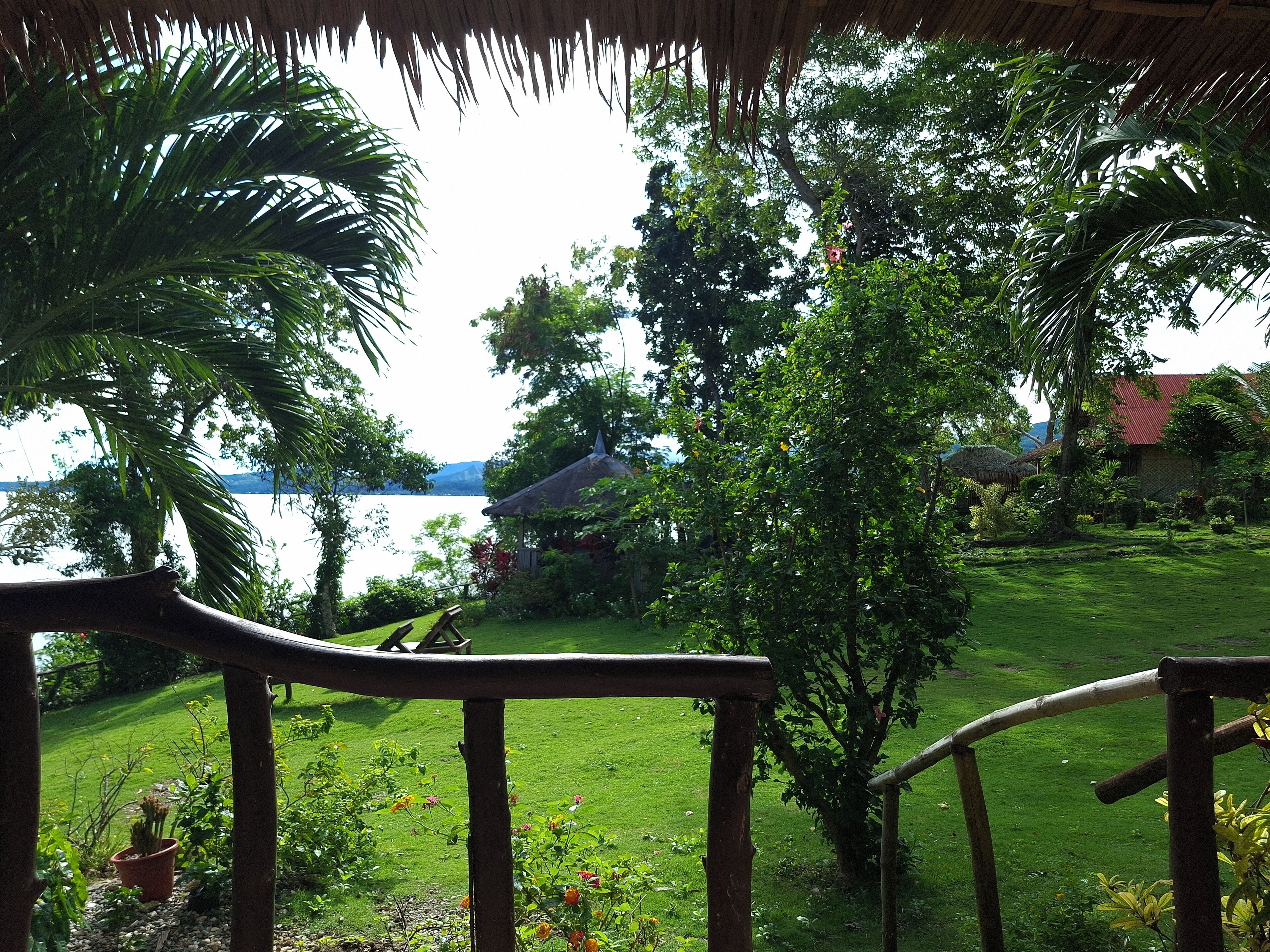 Vista de um deck de madeira com vista para um jardim exuberante, com uma casa e um corpo d'água ao fundo.