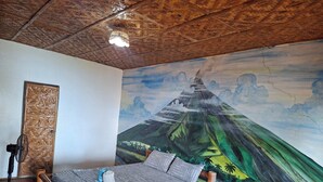 Une chambre avec un lit, un ventilateur, un plancher en bois et une fresque représentant un paysage de montagne.