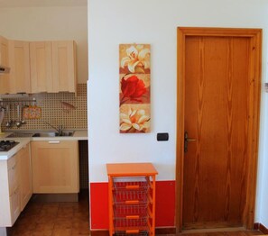 Una cocina con armarios de madera, estufa y lavamanos. En la pared hay un cuadro de flores y una puerta de madera.