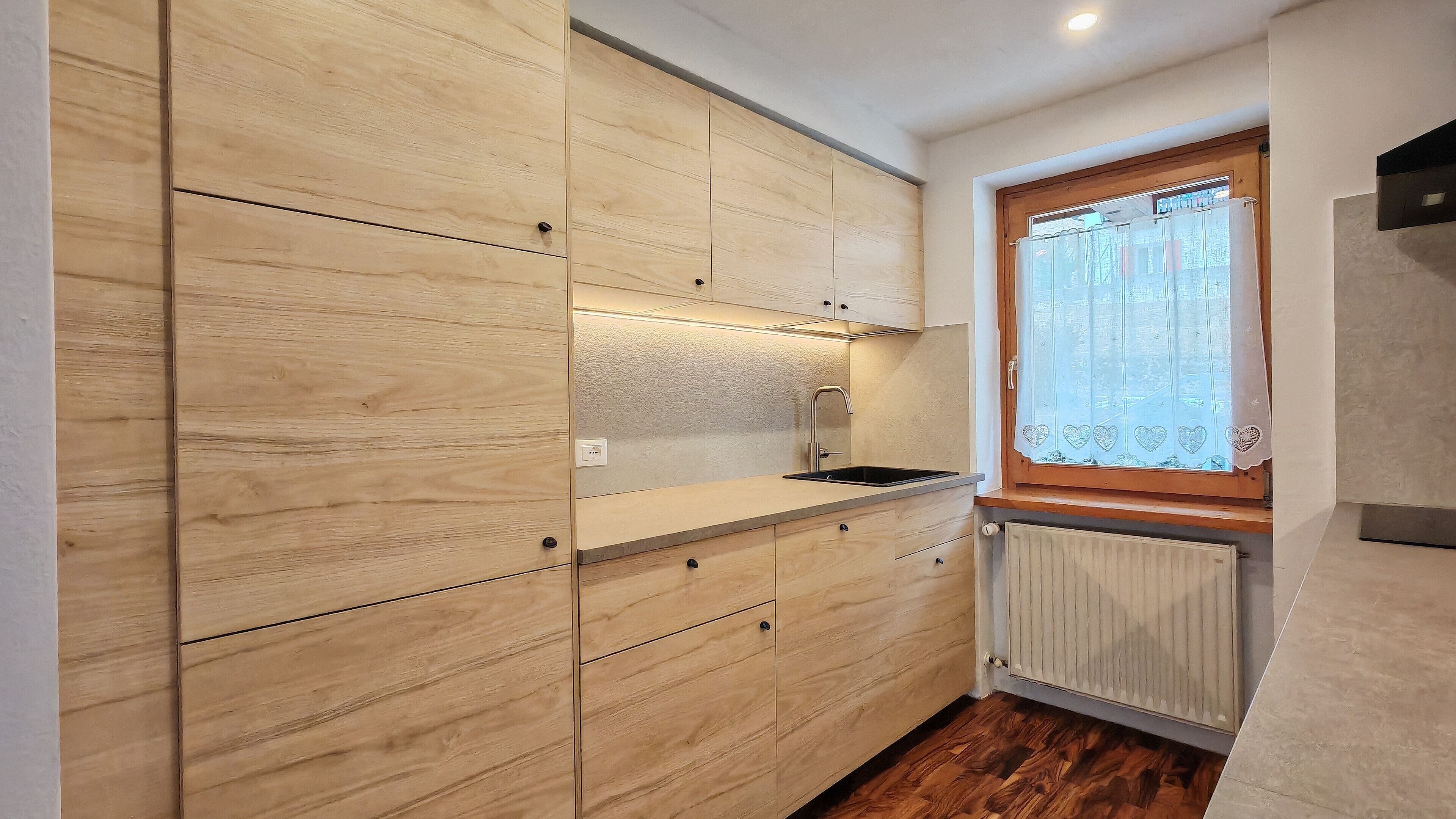 A compact kitchen with wooden cabinets, a window with a heart-patterned curtain, and a built-in oven.