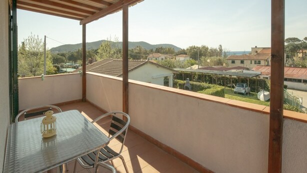Un balcon avec une table et des chaises en métal, d’où l’on peut admirer des maisons et des collines au loin.