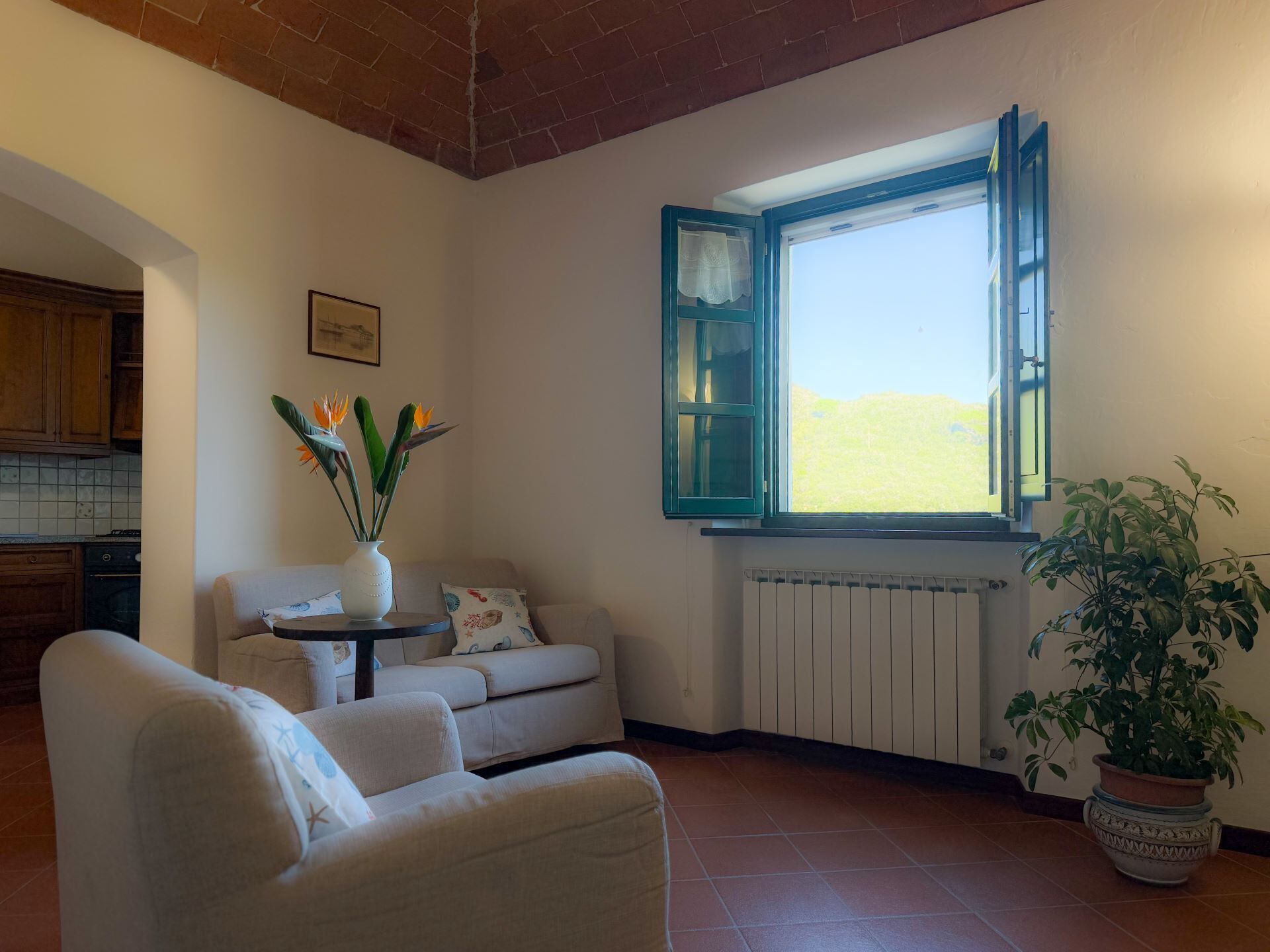 Uma sala de estar com um sofá, uma mesinha com um vaso de flores, uma planta em vaso e uma janela com vista para o exterior.
