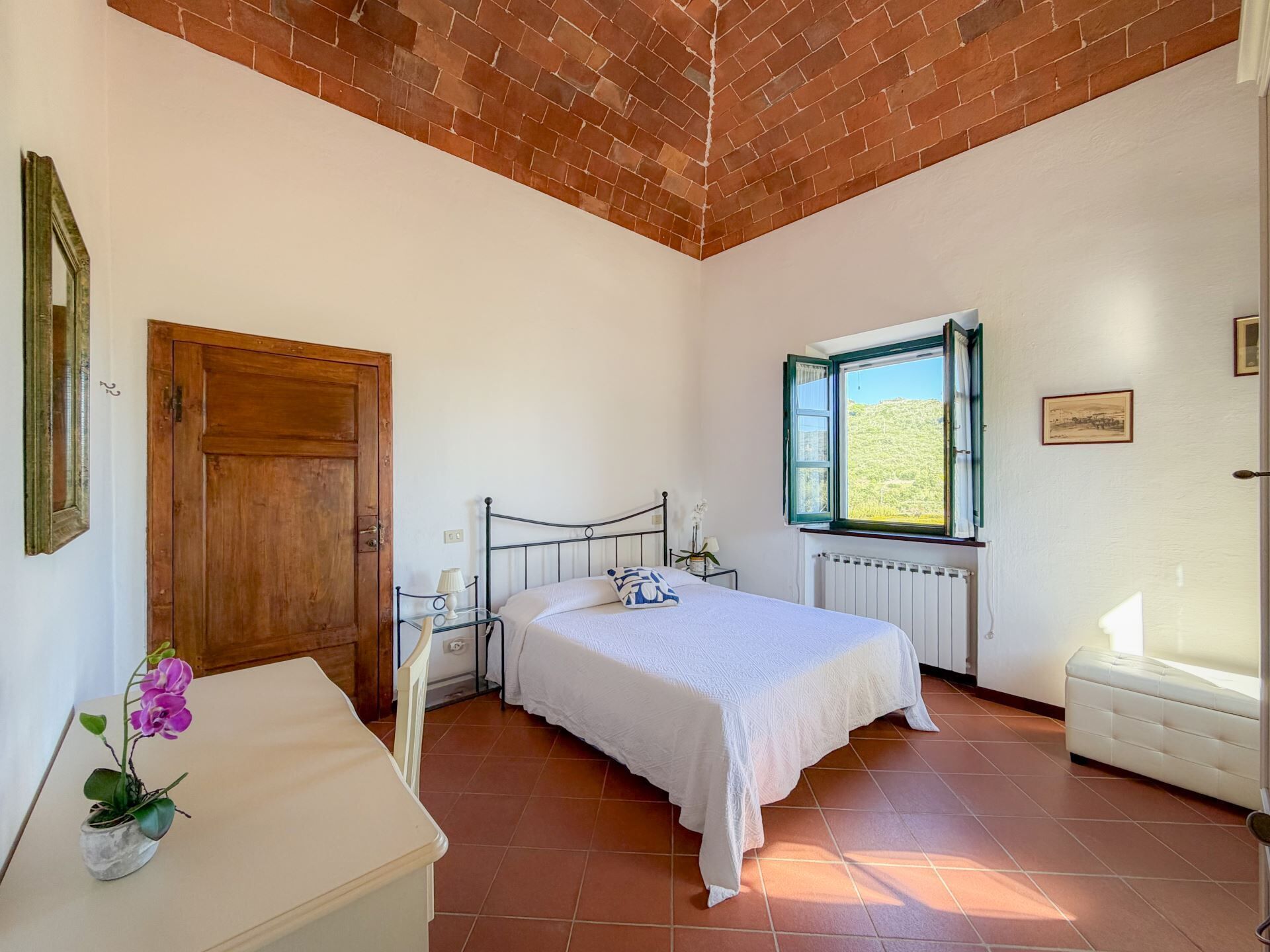 A bedroom with a bed, a wooden door, a window with a view, a radiator, and a small table with a lamp and flowers.