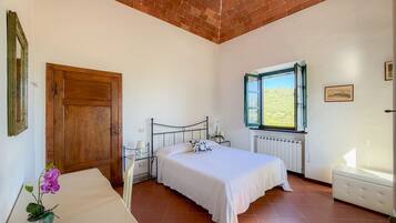 A bedroom with a bed, a wooden door, a window with a view, a radiator, and a small table with a lamp and flowers.