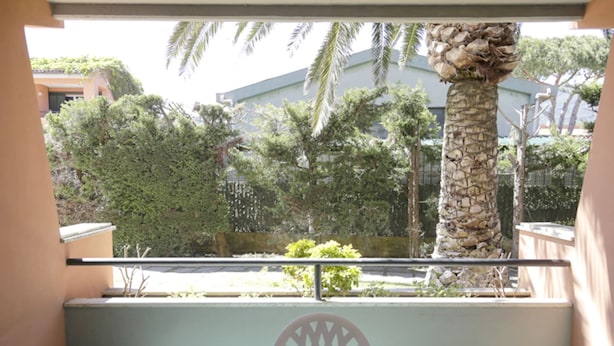 A balcony with a table and chairs, overlooking a garden with palm trees and a building in the background.