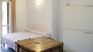 Et soveværelse med en seng, et bord og en stol. Der er et vindue med gardiner, en dør og en lampe på væggen.
