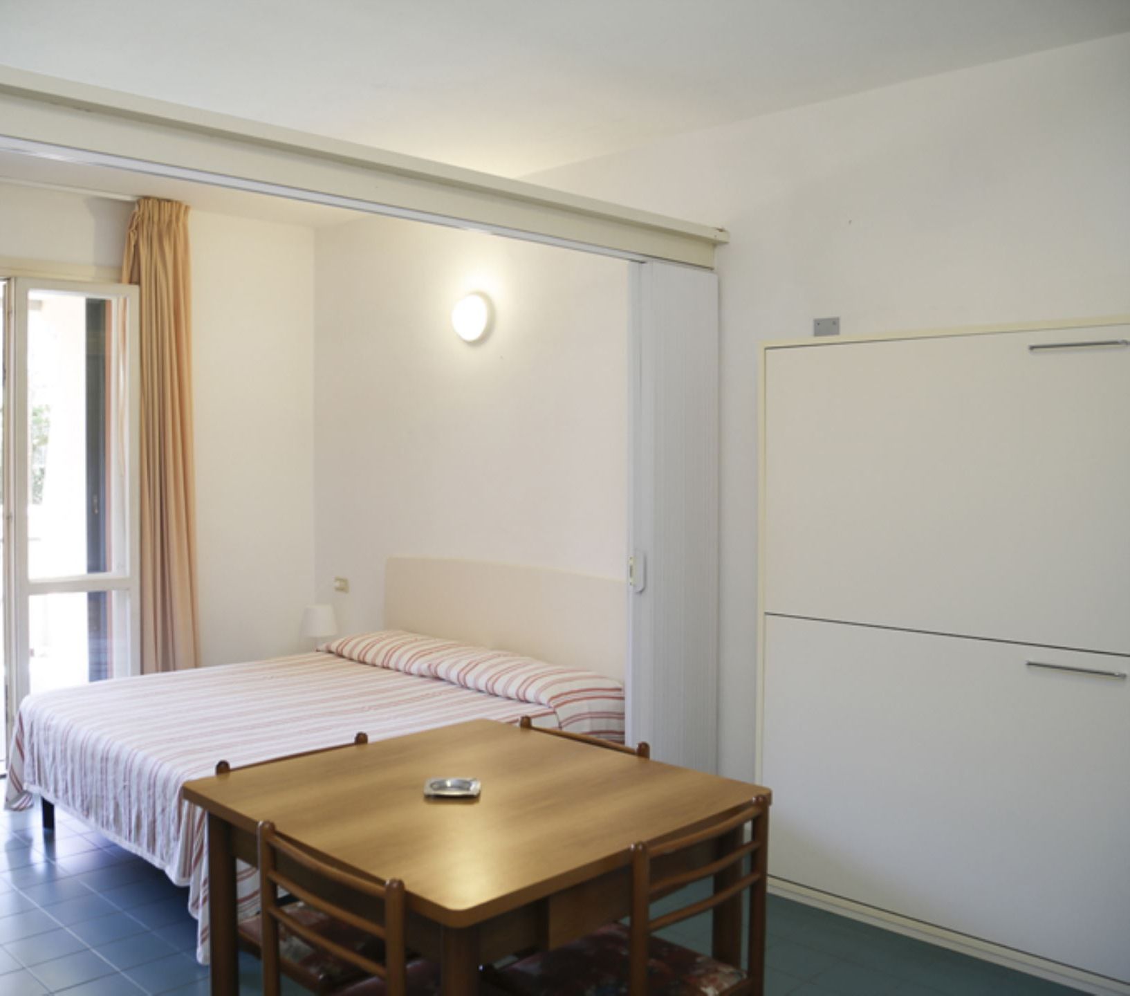 A bedroom with a bed, a table, and a chair. There is a window with curtains, a door, and a light fixture on the wall.