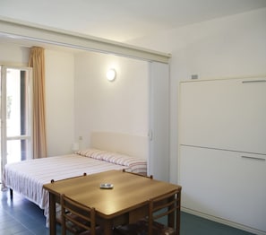 Une chambre avec un lit, une table et une chaise. Il y a une fenêtre avec des rideaux, une porte et un luminaire fixé au mur.