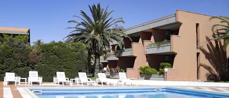 Una piscina con sillones blancos y un edificio con balcones.