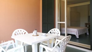 A small, private balcony with a white table and chairs, a coffee maker, and a bed with a striped bedspread visible through a sliding door. - The apartments, all studios, are simply and functionally furnished and all consist of a living room (Marina di Campo)