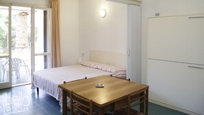 A bedroom with a bed, a table, and a chair. There is a window with curtains, a door, and a light fixture on the wall. - The apartments, all studios, are simply and functionally furnished and all consist of a living room (Marina di Campo)
