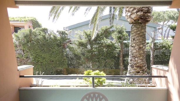 A balcony with a table and chairs, overlooking a garden with palm trees and a building in the background. - The apartments, all studios, are simply and functionally furnished and all consist of a living room (Marina di Campo)