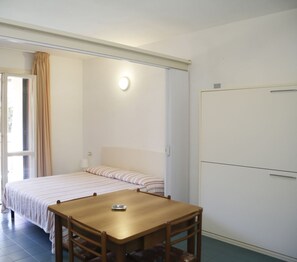 Une chambre avec un lit, une table et une chaise. Il y a une fenêtre avec des rideaux, une porte et un luminaire fixé au mur.