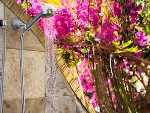 Ducha con manguera, pared de azulejos y flores rosadas al fondo.