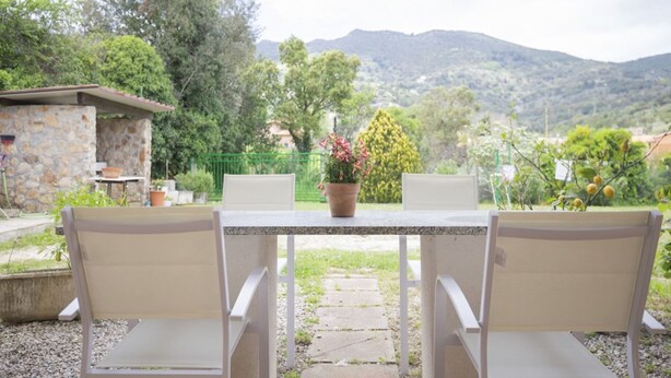 A patio with two white chairs and a table, surrounded by greenery and a stone building.