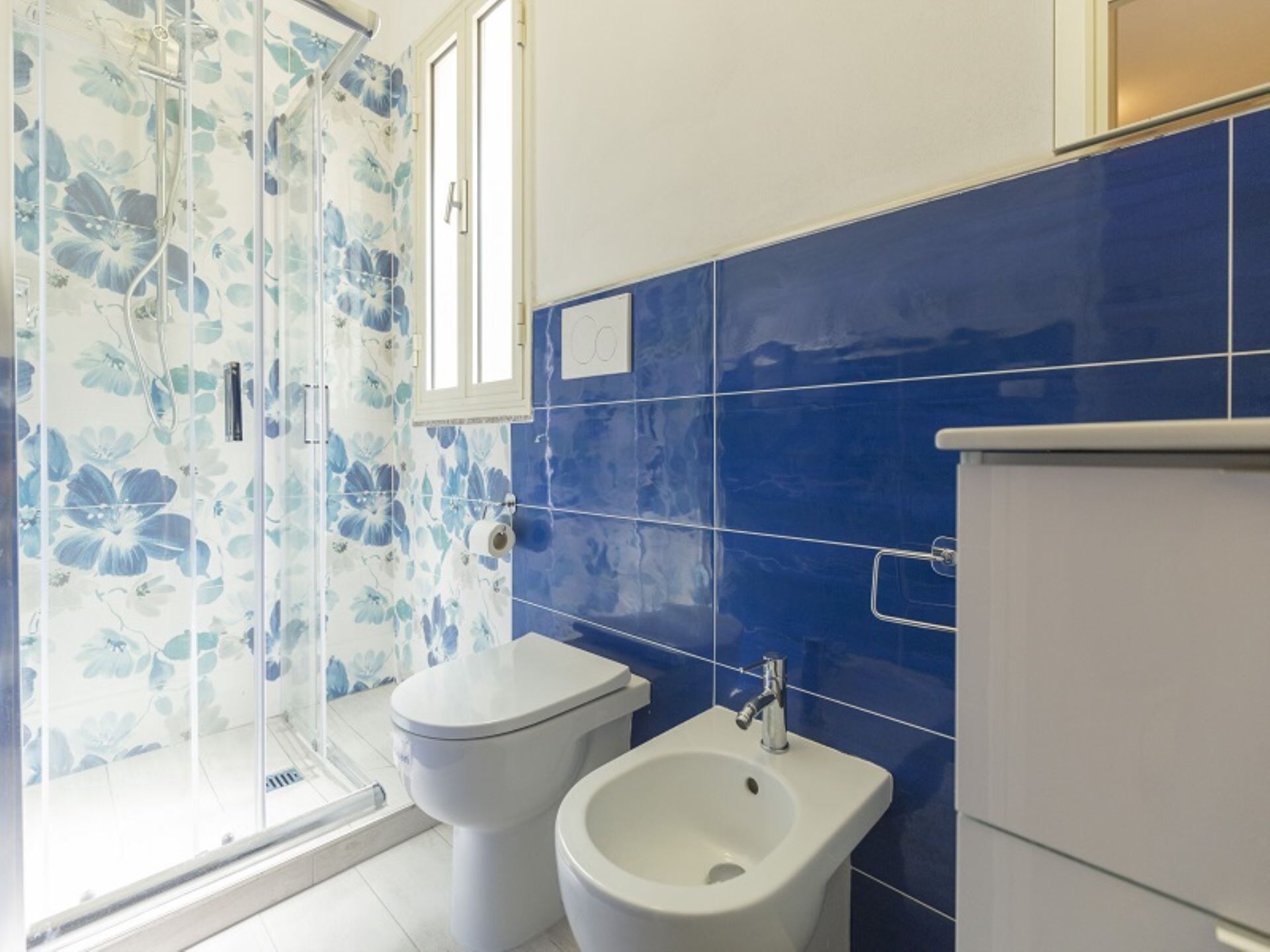 A bathroom with a shower enclosure, toilet, bidet, and a cabinet.
