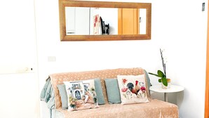 Ein gemütliches Zimmer mit einem beigefarbenen Sofa, gemusterten Kissen, einem kleinen runden Tisch mit einer Topfpflanze und einem großen Spiegel an der Wand.