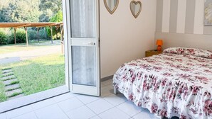 Ein Schlafzimmer mit Bett, Nachttisch, Lampe und Blick auf einen Garten durch eine geöffnete Tür.