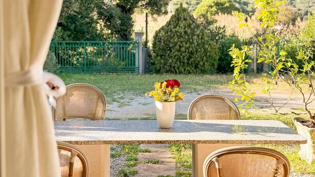 Ein Essbereich im Freien mit Korbstühlen und einem Tisch, eine Vase mit gelben Blumen sowie ein Blick auf einen grünen Zaun und Bäume im Hintergrund.
