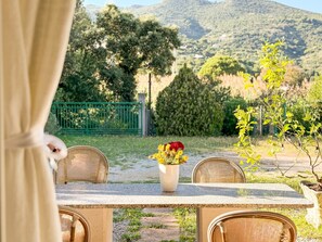 An outdoor dining area with wicker chairs and a table, a vase with yellow flowers, and a view of a green fence and trees in the background. - Within a private property, surrounded by a large garden, we offer our guests a spacious, fully equip (Rio Marina)