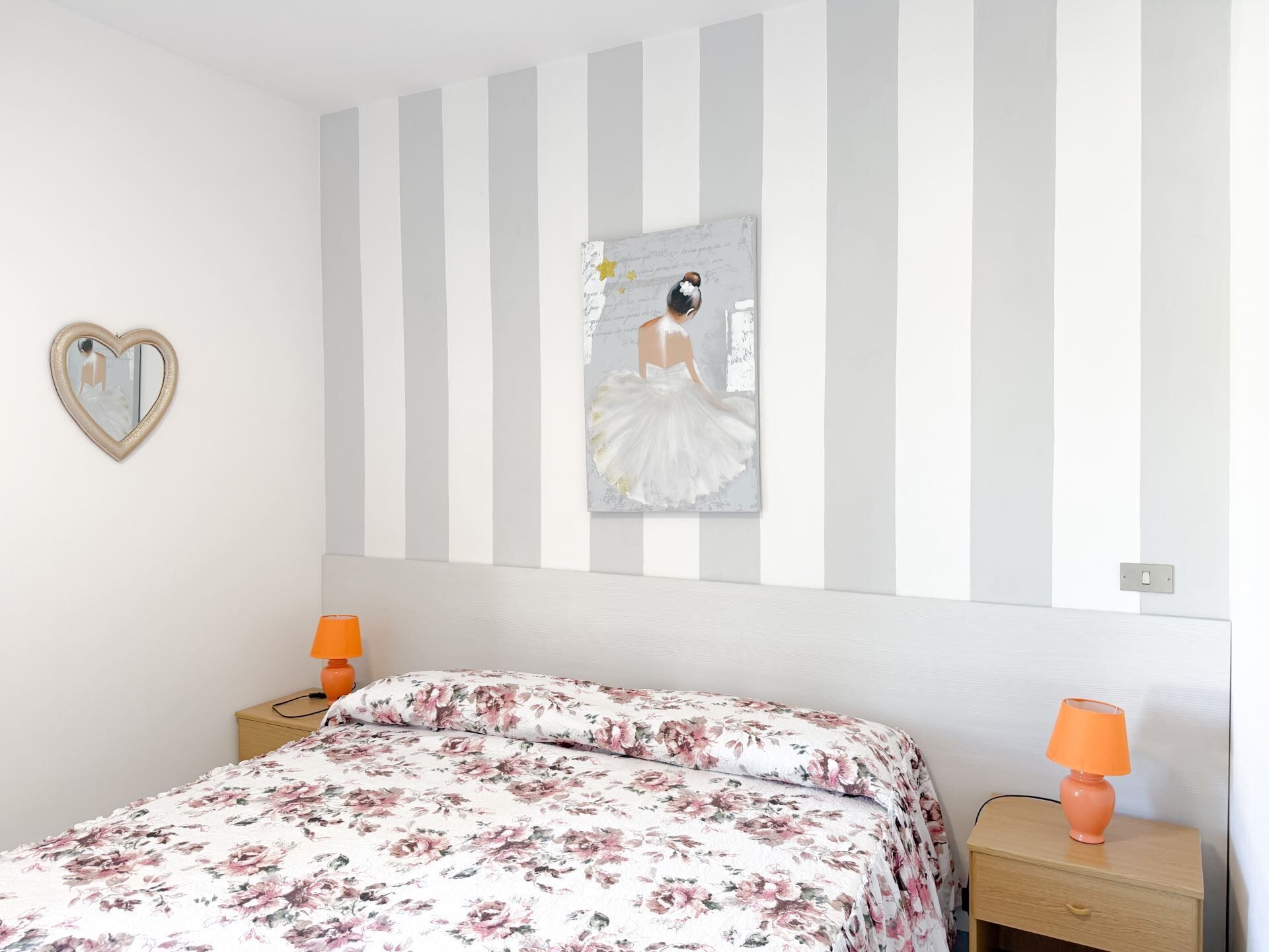 A bedroom with a bed, a heart-shaped mirror, a wall with a striped pattern, and a framed picture of a ballerina.