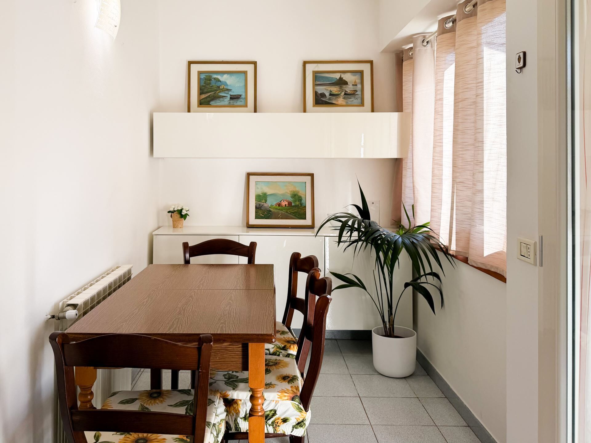 A dining area with a wooden table and chairs, a potted plant, and three framed paintings on the wall.