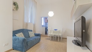 Un espacio habitable compacto con un sofá azul, una mesa de comedor con sillas, un televisor de pantalla plana y una zona de cocina al fondo.