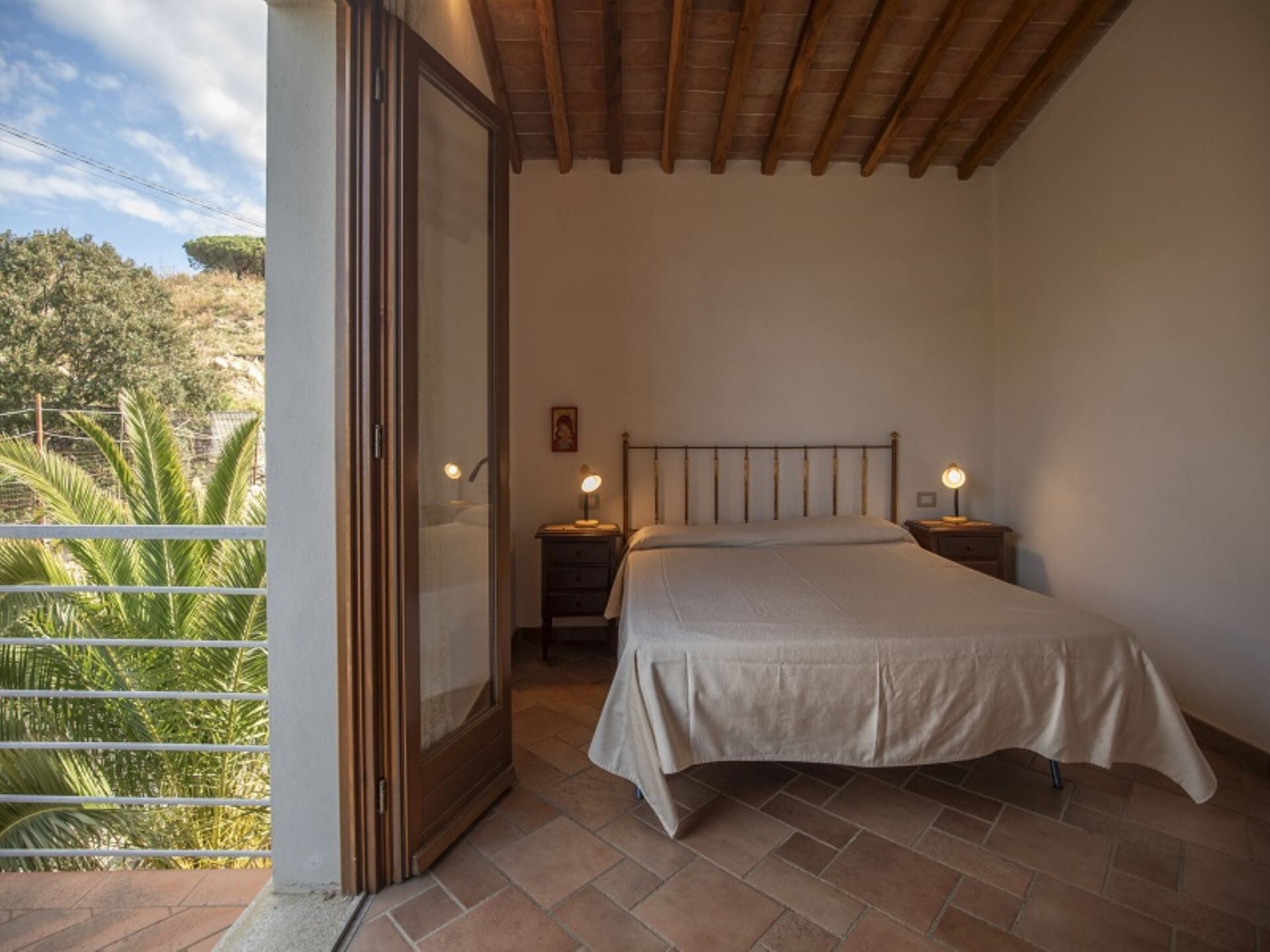 A bedroom with a bed, bedside tables, and a balcony with a view of greenery.