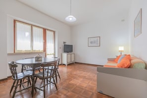 A room with a dining table, chairs, a television, a sofa, and a lamp.
