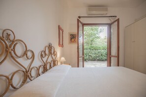 Ein Schlafzimmer mit einem Bett, einer Nachttischlampe, einem Bild an der Wand und einer offenen Tür, die in einen Garten führt.