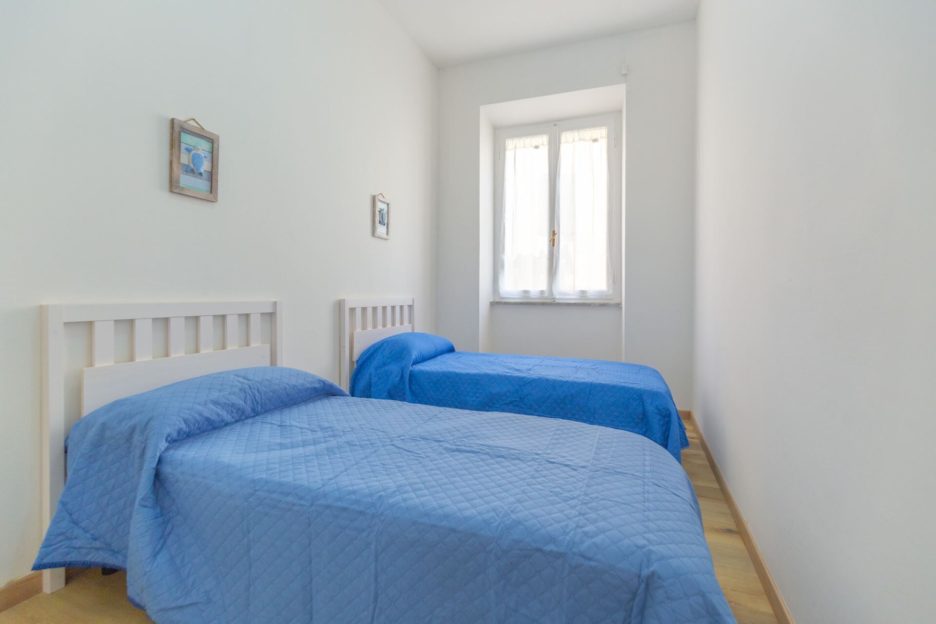 Een kamer met twee bedden, witte wanden, een raam met dunne gordijnen en een klein, ingelijst schilderij aan de muur.
