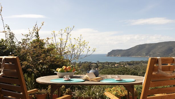 A round wooden table with a bowl of fruit, a teapot, and two cups, set against a scenic coastal backdrop. - It is located on the ground floor of a building consisting of three terraced houses. ' It consists o (Capoliveri)