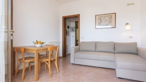 A living room with a beige sofa, a wooden dining table with chairs, and a bowl of fruit. - It is located on the ground floor of a building consisting of three terraced houses. ' It consists o (Capoliveri)