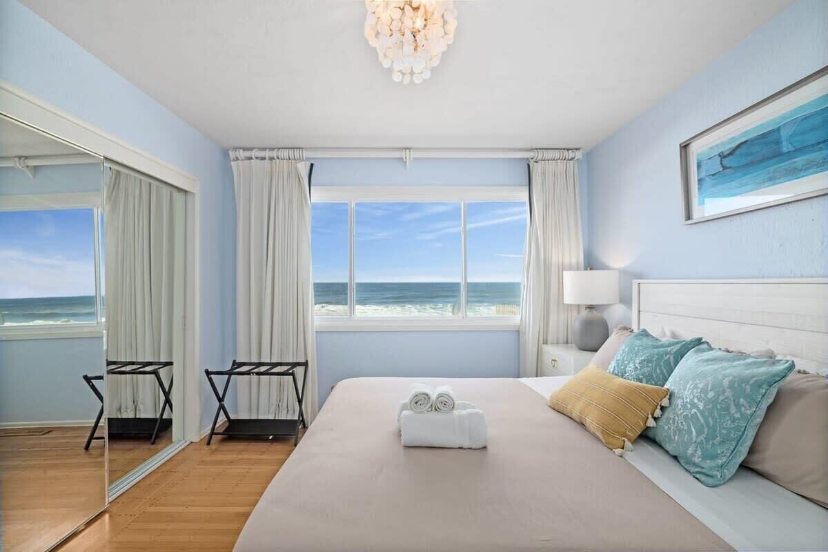 A bedroom with a bed, a window with a view of the beach, a chandelier, and a bedside table with a lamp.