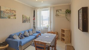 Ein gemütliches Wohnzimmer mit einem blauen Sofa, einem Esstisch mit gestreifter Tischdecke und einem Holzschrank. An den Wänden befinden sich Gemälde und rechts hängt eine Pflanze.