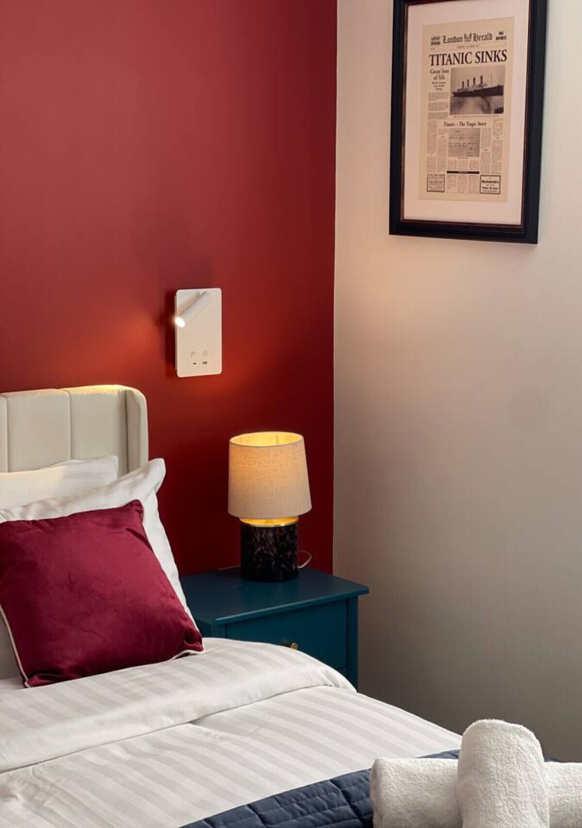 A hotel room with a bed, a nightstand with a lamp, a framed newspaper on the wall, and a red accent wall.