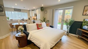 A modern bedroom with a large bed, a small side table, and a view of the kitchen and outdoor area. - Large New-Build 1 Bed House Near Science Park (Arbury)