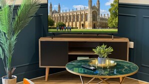 A living room with a large flat-screen TV displaying a building, a round glass coffee table, a sofa with cushions, and a potted plant. - Large New-Build 1 Bed House Near Science Park (Arbury)