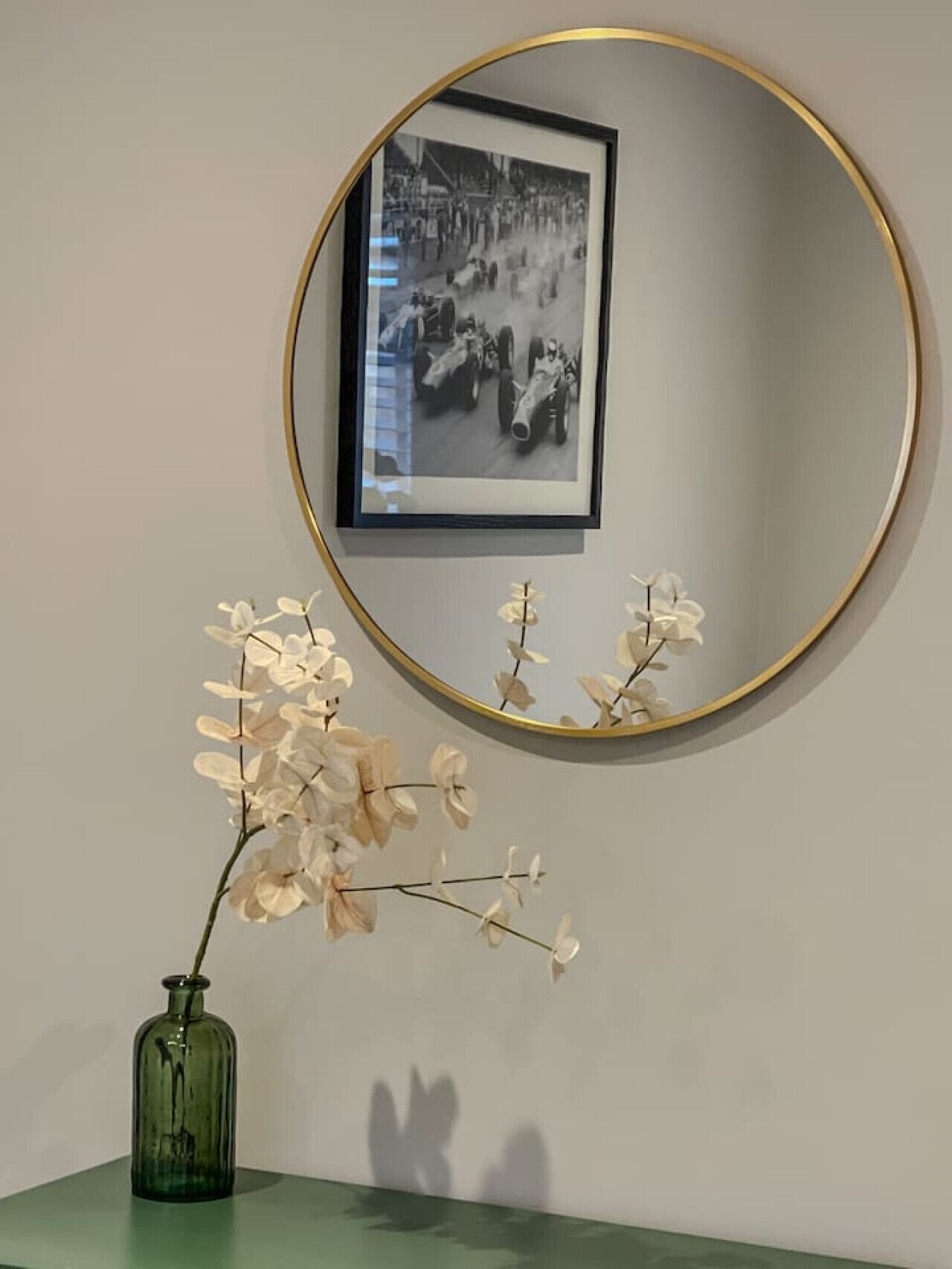 A framed black and white photograph of vintage racing cars on a wall, a green vase with white flowers on a table, and a round mirror with a gold frame.