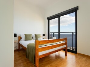 Un dormitorio con cama de madera, ropa de cama blanca y vista al mar a través de ventanas amplias.
