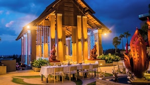 An outdoor dining area with a table set for two, surrounded by columns and string lights.