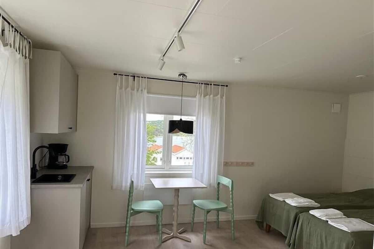 A small, clean room with a single bed, a table with two chairs, a kitchenette, and a window with curtains.