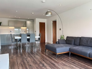 Ein modernes Wohnzimmer mit einem grauen Ecksofa, einem Essbereich mit weißem Tisch und grauen Stühlen sowie einer Küche im Hintergrund.