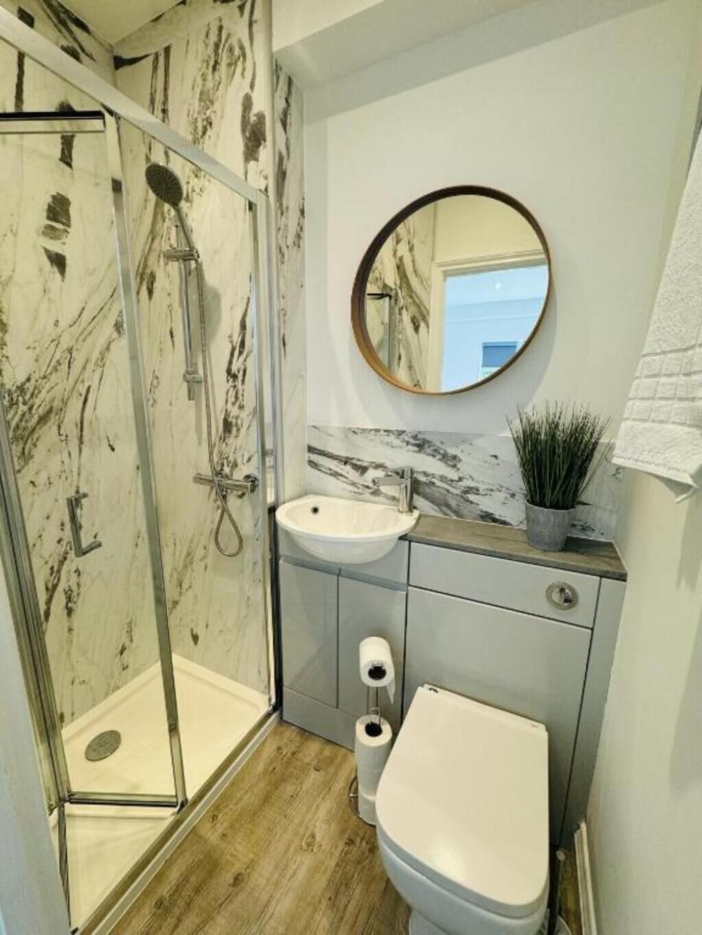 Un baño con cabina de ducha de vidrio, lavabo con encimera de mármol, inodoro y un espejo redondo en la pared.