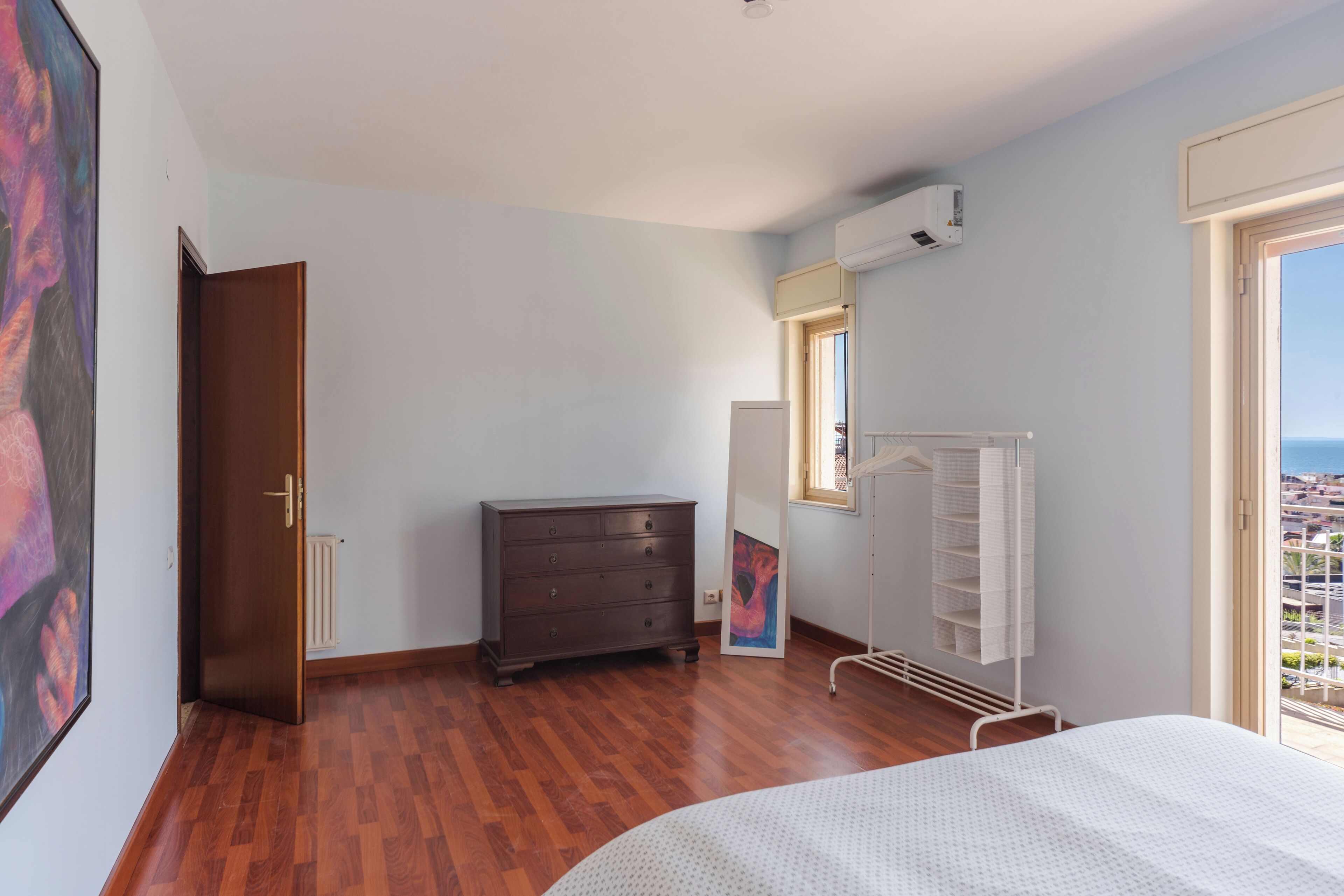 A bedroom with wooden flooring, a bed, a dresser, a wardrobe, and a window with a view.
