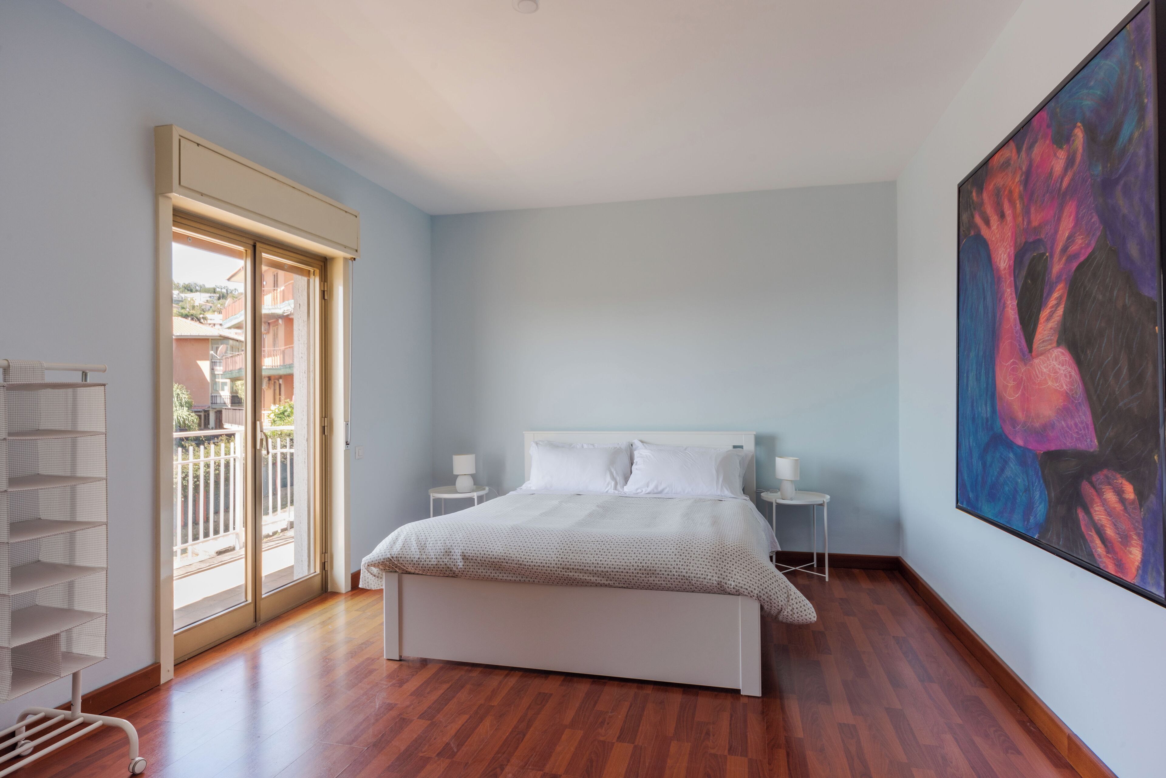 A bedroom with a bed, a nightstand, a wall-mounted painting, a sliding glass door, and a white shelving unit.
